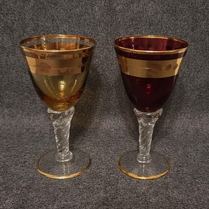 Op de afbeelding: Twee wijnglazen met gouden randen en bloemmotieven. Eén glas is amberkleurig, de andere dieprood. Beide hebben heldere, gedraaide stelen en goudgerande bases, tegen een grijze achtergrond.