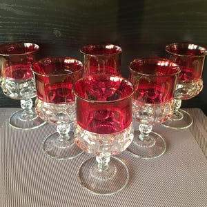 May include: Six ruby red wine glasses with clear, patterned bases and stems. The glasses have a classic design, with a vibrant red colour in the bowl and a clear, detailed base. The glasses are arranged on a striped surface.