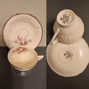 Puede incluir: Juego de taza y platillo de té color crema con detalles florales. La taza tiene un asa y se apoya en un soporte transparente. El platillo tiene un diseño floral en relieve y un borde dorado. Hecho en Inglaterra.