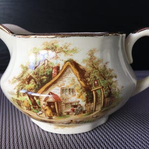 Puede incluir: Cremera de cerámica color crema con asa y boquilla, con una escena pastoral detallada. La obra de arte representa una cabaña con techo de paja, figuras y árboles, con ribetes dorados.