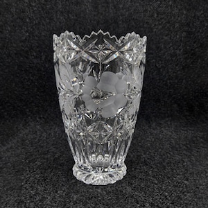 Peut inclure: Un vase en cristal transparent avec un motif floral détaillé. Le vase présente des fleurs et des feuilles gravées, avec un motif en étoile autour du bord et de la base. Le vase mesure environ 20 cm de haut.