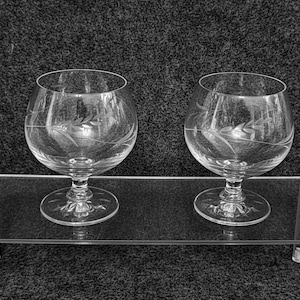 May include: Two clear brandy glasses with etched leaf patterns, presented on a clear acrylic shelf. The glasses feature a rounded bowl and a stemmed base. The background is a dark, textured surface.