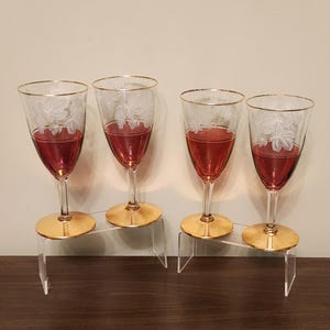 May include: Four stemmed wine glasses filled with red liquid. The clear glasses have gold rims and bases, with a white floral design etched on the upper portion. The glasses are displayed on clear acrylic stands.