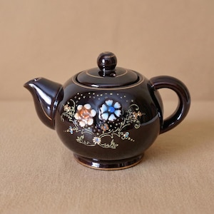 Può includere: Piccola teiera in ceramica marrone scuro con coperchio e manico. Decorata con motivi floreali blu, arancioni e bianchi. Rifinitura dorata sulla base e sul coperchio. Oggetto decorativo per il tè.