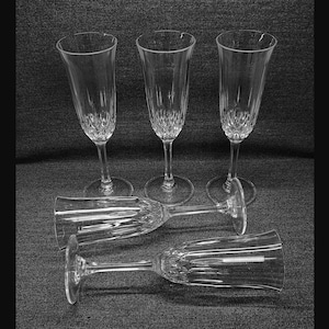 May include: A set of five clear glass champagne flutes featuring a tall, slender bowl with a faceted, diamond-cut pattern at the base. Three flutes stand upright on circular bases, while two flutes are positioned horizontally in the foreground.