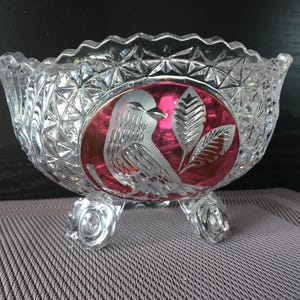 May include: A clear, footed crystal bowl with a scalloped rim. The bowl features a red oval with an etched bird and leaf design. The bowl has a diamond pattern and sits on three clear, decorative feet.