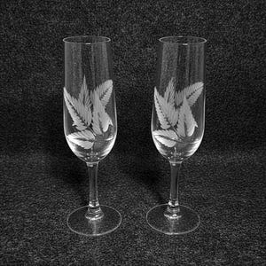 May include: Two clear champagne flutes with etched leaf designs. The flutes have a tall, slender shape with a round base and a long stem. The leaf design is etched into the glass, creating a delicate and elegant appearance.