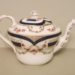 Könnte beinhalten: Weiße Porzellan-Teekanne mit Deckel, Henkel und Ausguss. Die Teekanne ist mit rosa Blumenmustern, goldenen Akzenten und marineblauen Bändern verziert. Ein kleiner goldener Knauf sitzt auf dem Deckel. Wahrscheinlich Vintage oder antik.