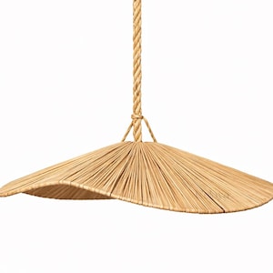 May include: A natural woven pendant light with a wide, gently curved shade. The shade is made of thin, light brown reeds radiating from a central point. It hangs from a thick, twisted rope, creating a rustic, organic aesthetic.