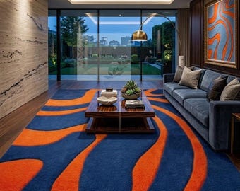 May include: A large rectangular area rug featuring a bold, wavy pattern in vibrant orange and deep blue. The rug covers a dark wood floor in a living room, positioned beneath a low-profile wooden coffee table with books and a small potted plant.