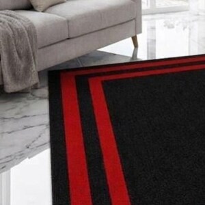 May include: A rectangular area rug featuring a solid black center field framed by two distinct, parallel red border stripes. The rug has a low-pile texture and is positioned on a white marble floor in front of a light gray sofa and large windows.