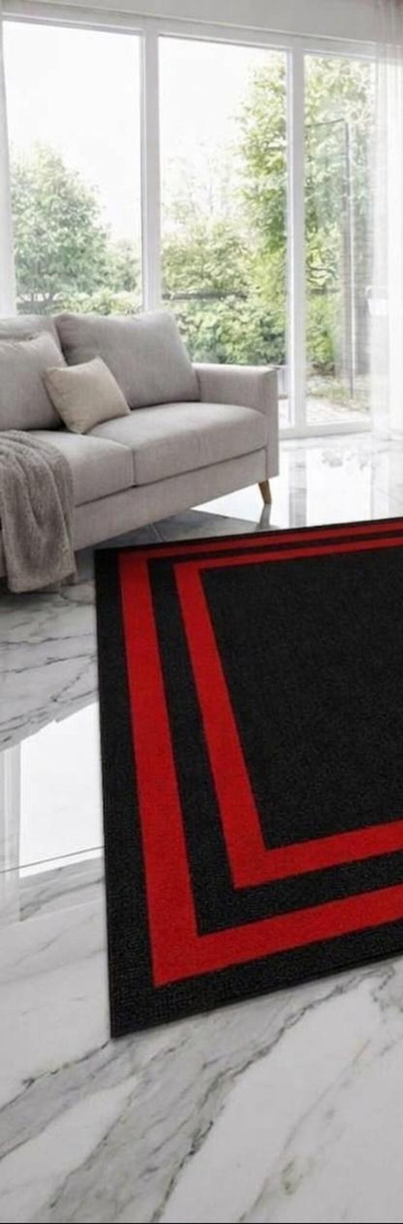 May include: A rectangular area rug featuring a solid black center field framed by two distinct, parallel red border stripes. The rug has a low-pile texture and is positioned on a white marble floor in front of a light gray sofa and large windows.