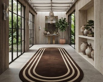May include: An elongated oval area rug featuring a dark brown centre surrounded by concentric light beige and brown stripes. The rug is positioned on a grey tiled floor in a hallway with wood-panelled walls, large glass windows, and pottery.