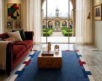 May include: A living room featuring a deep red velvet sofa, a rectangular wooden coffee table with a book and clock, and a large blue area rug with a geometric border in red and cream. A chandelier hangs above, and sheer curtains frame a view.