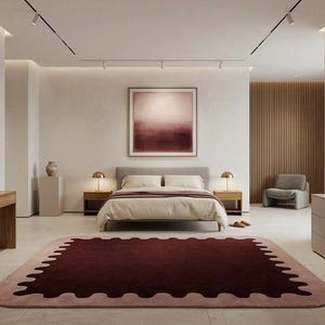 May include: A rectangular area rug featuring a deep burgundy center with a scalloped edge border in a lighter dusty pink tone. The rug is positioned in a bedroom with a bed, two wooden nightstands, gold lamps, and a wall-mounted abstract art piece.