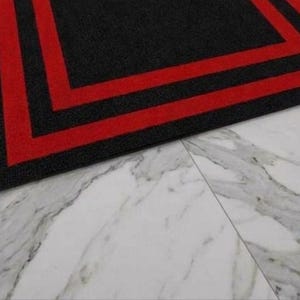May include: A rectangular area rug featuring a solid black center surrounded by two distinct, parallel red border stripes that frame the perimeter. The rug has a textured surface and is positioned on a white marble floor with grey veining.