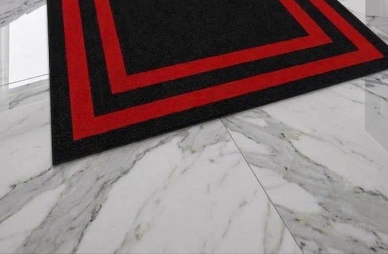 May include: A rectangular area rug featuring a solid black center surrounded by two distinct, parallel red border stripes that frame the perimeter. The rug has a textured surface and is positioned on a white marble floor with grey veining.