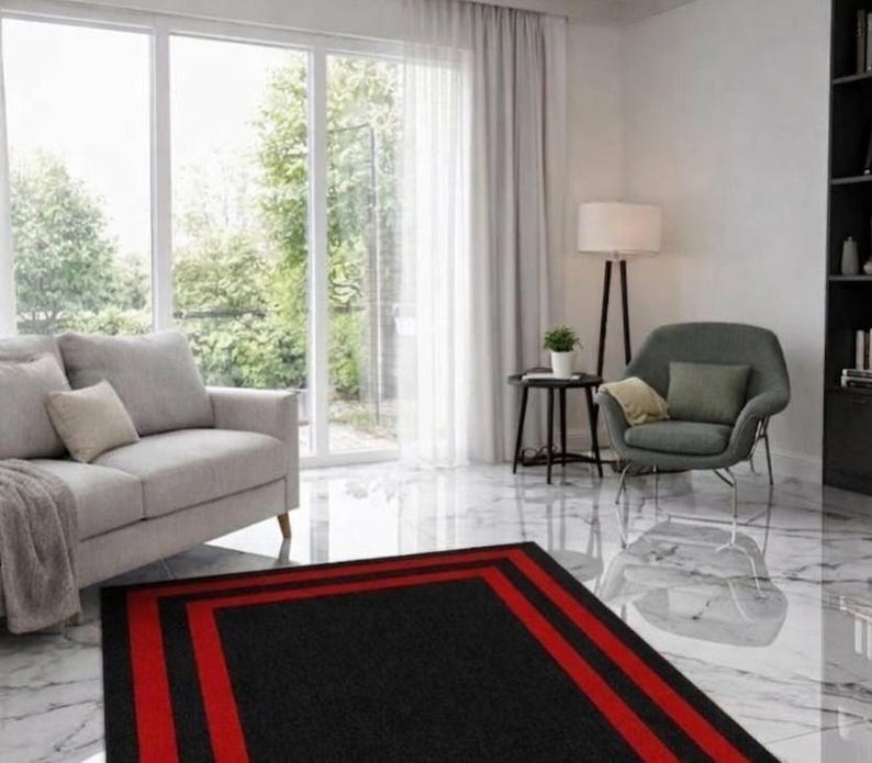 May include: A rectangular area rug featuring a solid black center framed by two bold red stripes and a black border. The rug is positioned on a white marble floor in a living room containing a gray sofa, a gray armchair, and a floor lamp.