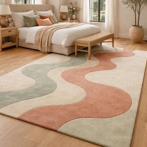 May include: A large, rectangular area rug with a wavy design in shades of beige, sage green, and coral. The rug is placed in a bedroom setting, with a bed, bench, and wooden flooring visible. The rug's dimensions are approximately 8 x 10 feet.