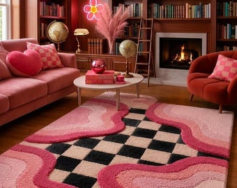 May include: A rectangular area rug featuring a central black and cream checkered pattern bordered by layered, wavy sections in various shades of pink. The rug has a textured, tufted surface and is positioned on a wooden floor in a library room.