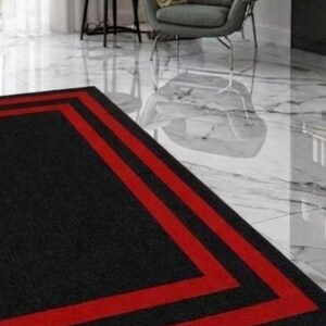 May include: A rectangular area rug featuring a solid black center bordered by two distinct, parallel red stripes that frame the perimeter. The rug is positioned on a white marble floor with grey veining, set within a room containing a grey armchair.