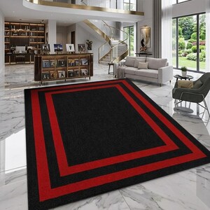 May include: A rectangular area rug featuring a solid black center field framed by two distinct, bold red rectangular borders. The rug has a low-pile texture and is positioned on a white marble floor within a large, open-concept living space.