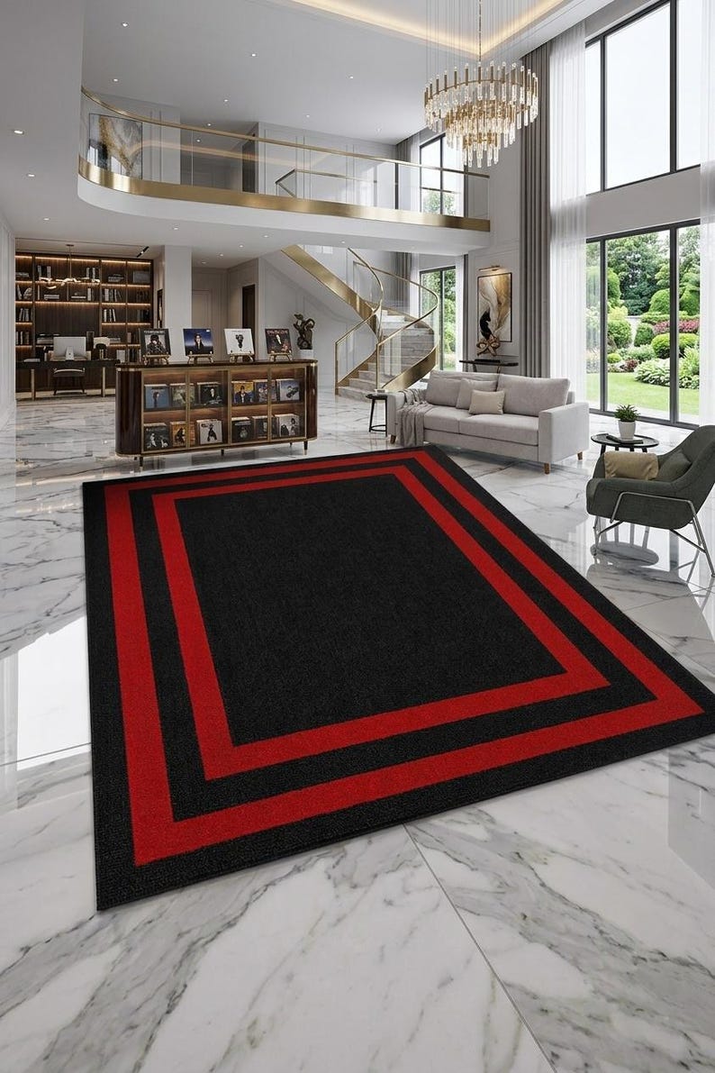 May include: A rectangular area rug featuring a solid black center field framed by two distinct, bold red rectangular borders. The rug has a low-pile texture and is positioned on a white marble floor within a large, open-concept living space.