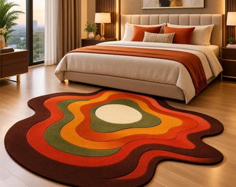 May include: An irregular, organic-shaped area rug featuring concentric, wavy bands of colour in shades of dark brown, bright orange, muted green, and a central cream-coloured circle. The rug lies flat on a light-toned wooden floor in a bedroom.