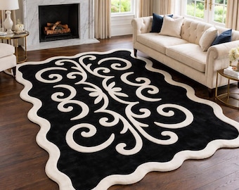 May include: A rectangular area rug featuring a solid black field with a thick, cream-coloured scalloped border and a central cream-coloured symmetrical scrollwork pattern. The rug is positioned on a dark brown hardwood floor in a living room.