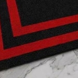 May include: A rectangular floor mat featuring a textured black surface with two bold, parallel red stripes forming a right-angle corner design. The mat has a dense, low-pile construction and clean, straight edges that define its geometric pattern.
