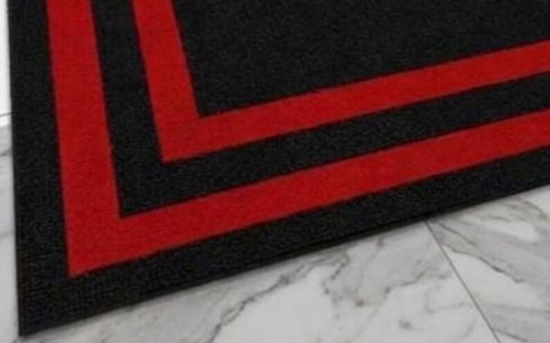 May include: A rectangular floor mat featuring a textured black surface with two bold, parallel red stripes forming a right-angle corner design. The mat has a dense, low-pile construction and clean, straight edges that define its geometric pattern.
