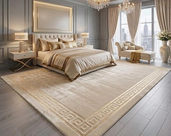 May include: A large rectangular area rug featuring a solid beige centre and a wide border with a repeating gold Greek key pattern. The rug is placed on a light grey wood floor in a bedroom with a tufted headboard bed and two crystal chandeliers.