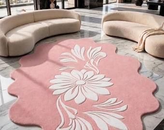 May include: A large, pink, organic-shaped area rug featuring a prominent white floral motif with stylised petals and leaves. The rug has a soft, textured surface and a scalloped edge, creating a distinct contrast against the light pink background.