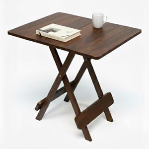 May include: A dark brown, folding wooden table with a square top. A white mug and a book are on the table. The table's legs are in an X-shape, and the table is approximately 24 inches wide.