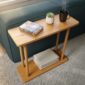 May include: A light brown wooden side table with a rectangular top and a lower shelf. A book, a small potted succulent, and a black mug are on the table. A folded grey blanket rests on the shelf. The table is next to a teal sofa.