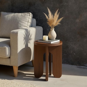 Puede incluir: Una pequeña mesa auxiliar redonda de madera marrón con un diseño de tres patas. La mesa sostiene libros, una vela encendida y un jarrón blanco con hierba ornamental seca. Un sofá beige está en el fondo. La mesa mide unos 40 cm de diámetro.