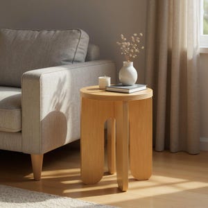 May include: A light-colored wooden side table with a round top and three curved legs. A small white vase with dried flowers, a candle, and a book sit on top. The table is approximately 18 inches tall.