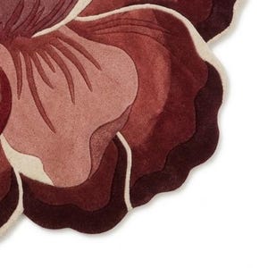 May include: A tufted rug shaped like a large flower with layered petals in shades of deep burgundy, dark red, and dusty rose. The petal edges are outlined with a thin, cream-colored border, creating a distinct contrast against the dark red tones.