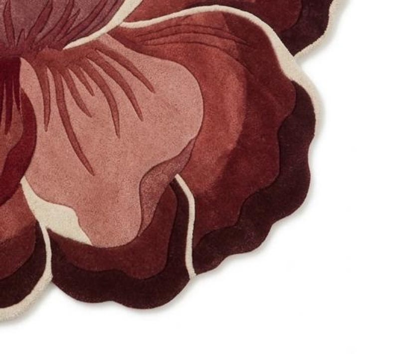 May include: A tufted rug shaped like a large flower with layered petals in shades of deep burgundy, dark red, and dusty rose. The petal edges are outlined with a thin, cream-colored border, creating a distinct contrast against the dark red tones.