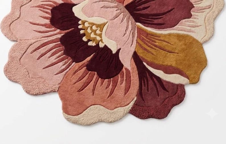 May include: A tufted area rug shaped like a large blooming flower with scalloped edges. The design features layered petals in shades of deep burgundy, dusty rose, soft pink, mustard yellow, and cream, creating a textured floral pattern.
