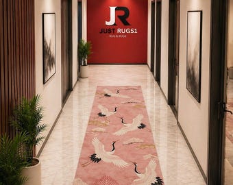 May include: A pink runner rug with a crane design in a hallway. The rug features white cranes with black accents and gold clouds. The hallway has white walls, dark doors, and artwork. The wall at the end of the hallway is red with the text "JUST RUGS1".