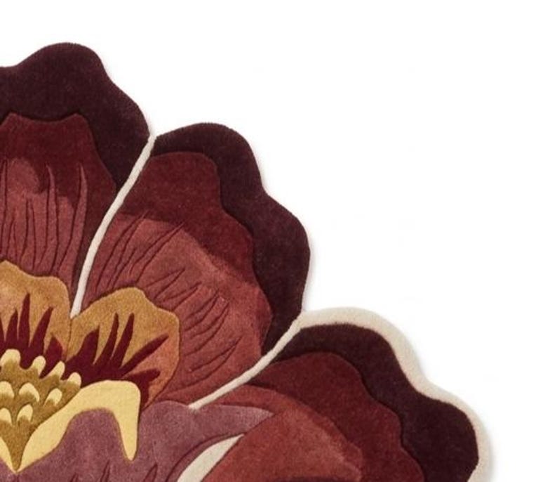 May include: A tufted rug shaped like a large flower with layered petals in shades of deep burgundy, maroon, and muted mauve. The center features golden yellow and light cream accents, with a distinct cream-colored border outlining the petal edges.