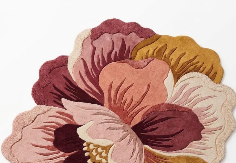 May include: A tufted rug shaped like a large blooming flower with layered petals in shades of deep burgundy, dusty rose, soft pink, cream, and mustard yellow. The rug features a textured surface with scalloped edges and intricate petal detailing.