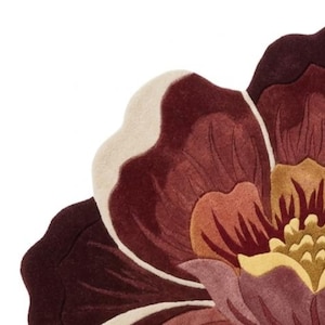 May include: A tufted rug shaped like a large flower with layered petals in shades of deep burgundy, muted mauve, soft cream, and golden yellow. The design features curved, scalloped edges and a dense, textured pile surface with distinct color sections.