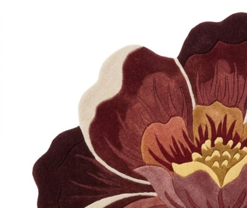 May include: A tufted rug shaped like a large flower with layered petals in shades of deep burgundy, muted mauve, soft cream, and golden yellow. The design features curved, scalloped edges and a dense, textured pile surface with distinct color sections.
