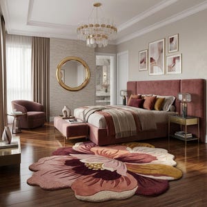 May include: A large, flower-shaped area rug featuring layered petals in shades of deep burgundy, dusty rose, and muted gold. The rug is placed on a dark wood floor in a bedroom with a velvet bed frame, a round gold mirror, and a crystal chandelier.
