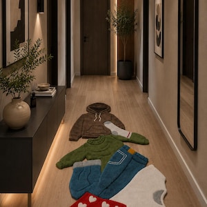 Può includere: Un corridoio con una selezione di originali tappeti a forma di capi d'abbigliamento. Tra questi, una felpa con cappuccio marrone, un maglione verde, jeans blu e pantaloncini rossi con cuori bianchi, su un pavimento in legno.