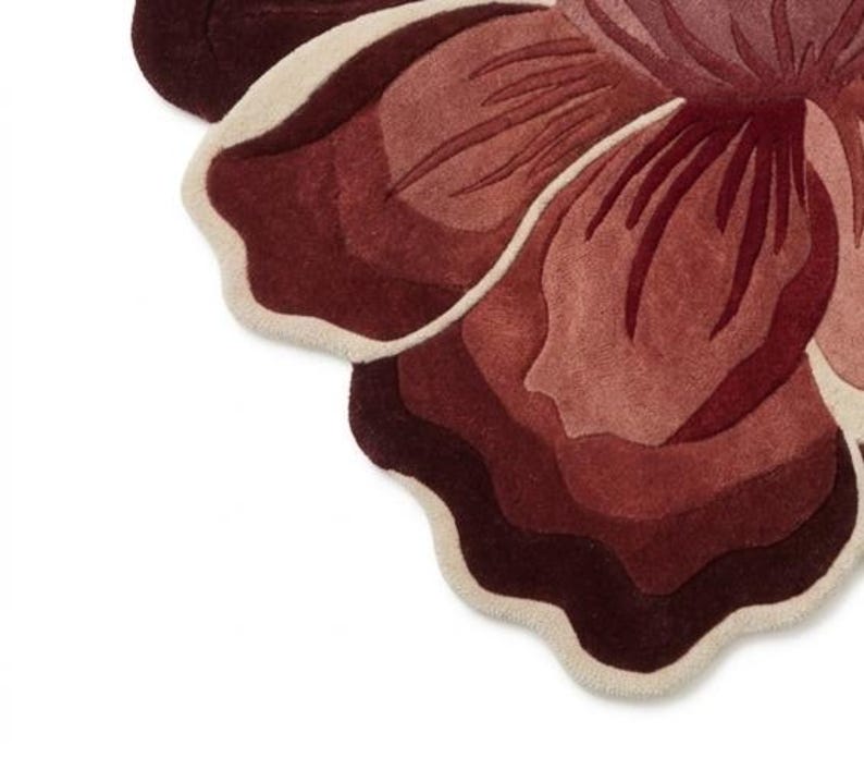 May include: A tufted rug shaped like a large flower with layered petals in shades of deep burgundy, dark red, and muted rose. The edges of the petals are outlined with a thick, cream-colored border, creating a distinct contrast against the dark tones.