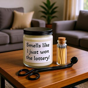 May include: A candle in a glass jar with a black lid and the text "Smells like I just won the lottery!". A small glass bottle with a cork stopper and matches, and a black candle wick trimmer are on a wooden table.