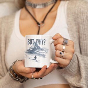 May include: White ceramic mug featuring the text "BUT WHY?" above a mountain scene and a penguin. The mountain is illustrated in blue and grey. The mug has a white handle.